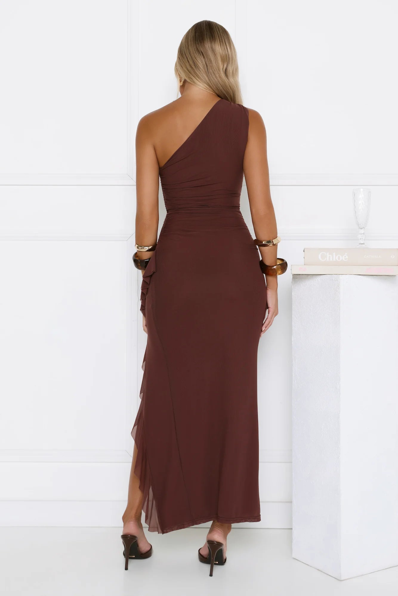 Exquisite chocolate brown one-shoulder maxi dress with beautifully tailored ruched waist detailing, a graceful ruffled high slit, and charming button accents on the shoulder.
